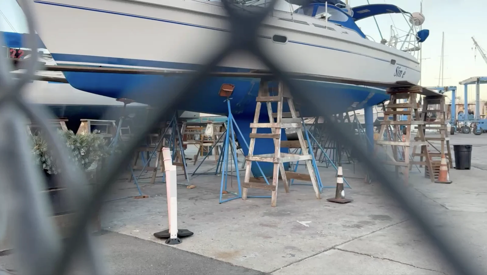 Sailboat resting on stands in a boatyard, seen through an out-of-focus fence.
