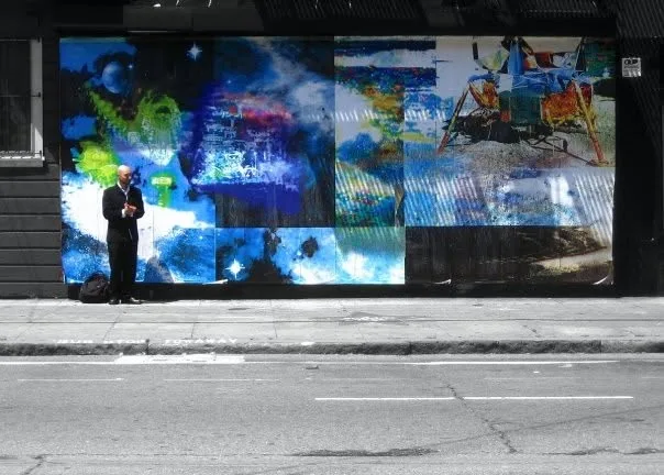 A person stands on a sidewalk in front of a large mural of space, planets, and a lunar lander.
